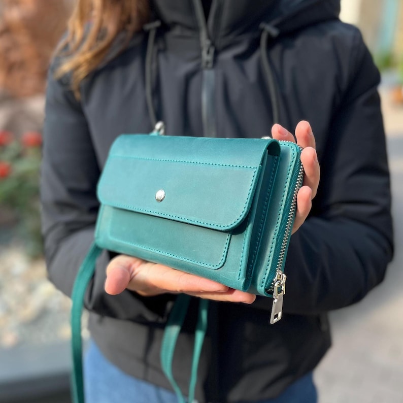 May include: A teal leather crossbody bag featuring a rectangular flap with a single silver-toned snap button closure, a side zipper compartment with a silver metal pull, and a matching thin leather shoulder strap for carrying.