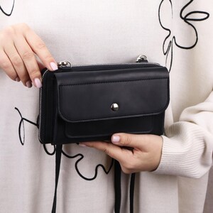 May include: A black leather crossbody bag featuring a rectangular silhouette, a front flap pocket with a silver-tone snap button closure, and a top zipper. The bag includes a thin black shoulder strap and silver-tone metal hardware attachments.