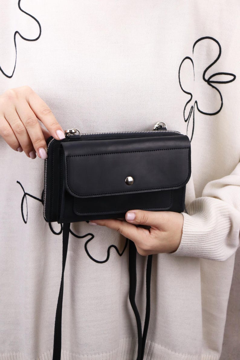 May include: A black leather crossbody bag featuring a rectangular silhouette, a front flap pocket with a silver-tone snap button closure, and a top zipper. The bag includes a thin black shoulder strap and silver-tone metal hardware attachments.