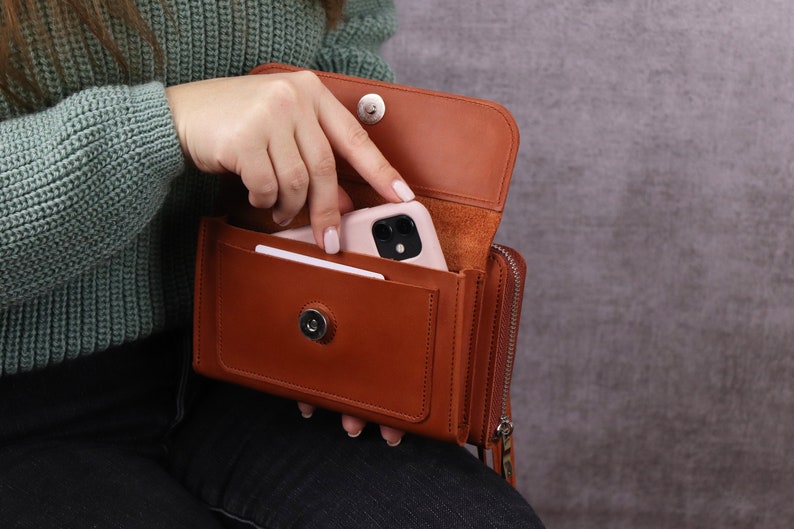 May include: A tan leather wallet with a magnetic snap closure, featuring an external pocket containing a light pink smartphone and a white card. The wallet has a side zipper compartment and a smooth, structured finish with visible stitching.