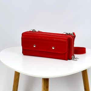 Puede incluir: Un bolso bandolera rojo brillante con forma rectangular. El bolso tiene un cierre de solapa con dos cierres a presión plateados y un compartimento con cremallera. Tiene una correa larga y ajustable y está sobre una mesa blanca.