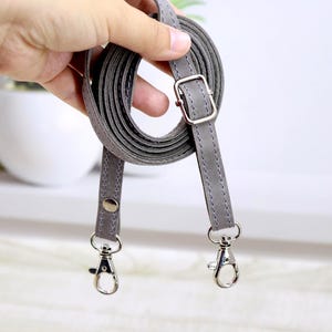 May include: A gray crossbody bag strap with silver-colored metal hardware. The strap is made of a smooth, faux leather material and features a buckle for adjusting the length. The strap has two silver-colored swivel clasps.