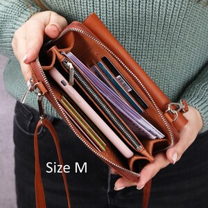May include: A tan leather crossbody wallet with a zipper closure, featuring multiple interior compartments, card slots, and a light pink smartphone. The interior contains paper currency and cards. The text Size M is printed on the lower left.