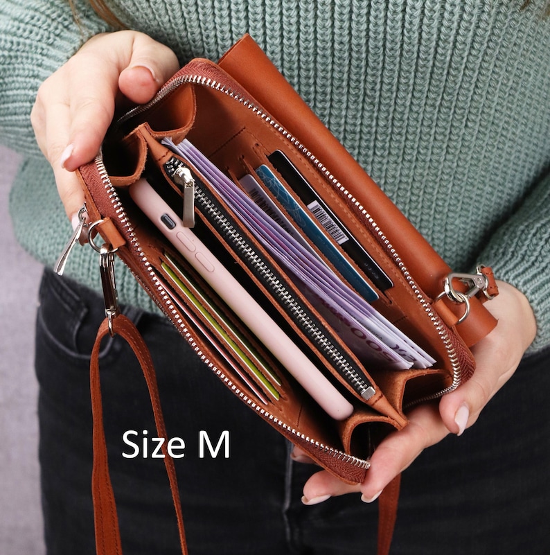 May include: A tan leather crossbody wallet with a zipper closure, featuring multiple interior compartments, card slots, and a light pink smartphone. The interior contains paper currency and cards. The text Size M is printed on the lower left.