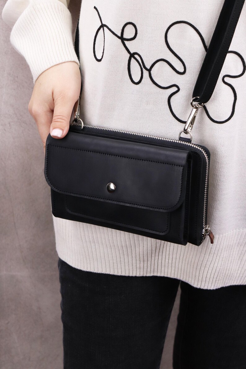 May include: A black rectangular crossbody bag featuring a front flap with a silver-toned snap button closure, a main zippered compartment, and a detachable black shoulder strap with silver metal hardware clips attached to the sides of the bag.