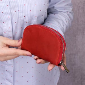 Może przedstawiać: Jasnoczerwony, półokrągły portfel ze srebrnym zamkiem błyskawicznym. Portfel jest prezentowany na tle jasnoniebieskiej koszuli z małymi czerwonymi wzorami serc.