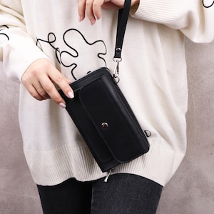 May include: A black rectangular wristlet wallet featuring a front flap pocket with a silver-toned snap button closure, a full-length zipper along the top, and a detachable black wrist strap attached to a silver metal ring on the side of the wallet.