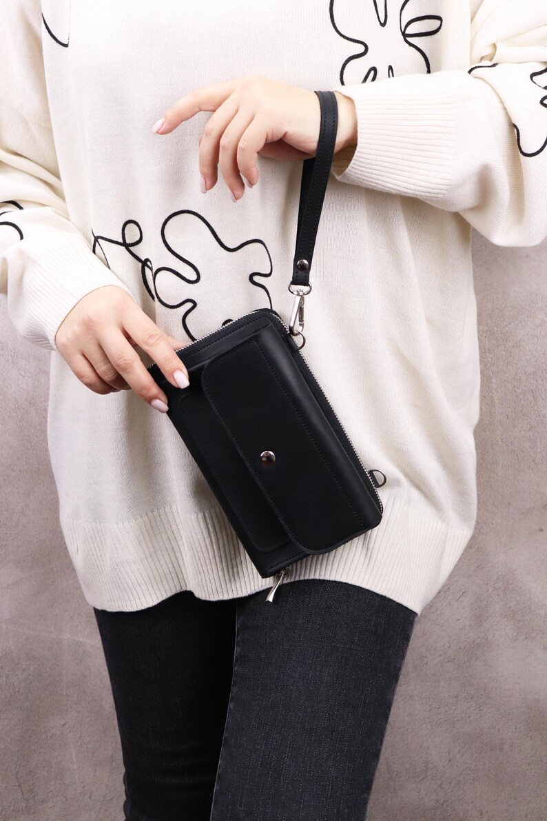 May include: A black rectangular wristlet wallet featuring a front flap pocket with a silver-toned snap button closure, a full-length zipper along the top, and a detachable black wrist strap attached to a silver metal ring on the side of the wallet.