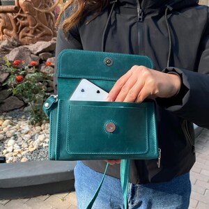 May include: A dark teal leather crossbody bag with a rectangular flap, a silver magnetic snap closure, and a side zipper. The bag features a front pocket containing a white rectangular object with black heart icons, and a thin matching leather strap.