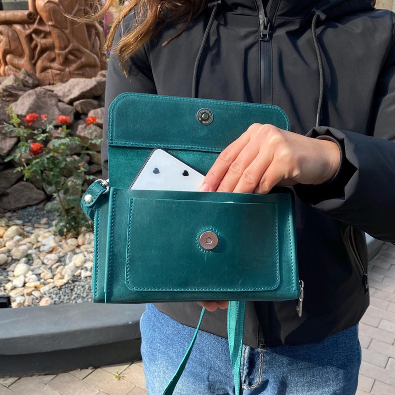 May include: A dark teal leather crossbody bag with a rectangular flap, a silver magnetic snap closure, and a side zipper. The bag features a front pocket containing a white rectangular object with black heart icons, and a thin matching leather strap.