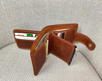 May include: A tan leather bifold wallet featuring multiple interior card slots, a central bill compartment, and a side zippered pocket. The wallet includes a snap button closure tab on the right side and contains several credit cards and identification.
