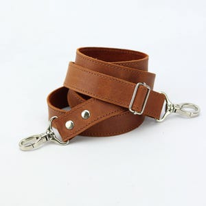 May include: A rolled, brown leather strap with silver-colored metal hardware. The strap has a buckle and two swivel clasps. The leather is a warm, medium brown color. The hardware is shiny and new.