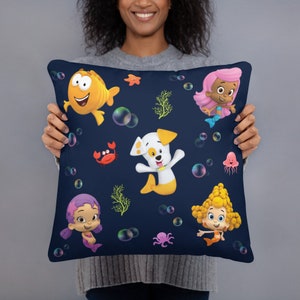 Op de afbeelding: Een blauw kussen met een kleurrijke onderwater scène met cartoonfiguren uit de show Bubble Guppies. De personages omvatten een gele vis, een witte hond, een roze zeemeermin en twee paarse zeemeerminnen. Het kussen is versierd met bubbels, zeewier en andere oceaan-thema elementen.