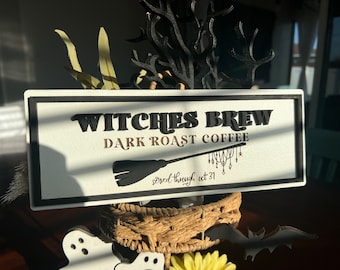 May include: A white sign with black lettering that reads "Witches Brew Dark Roast Coffee" with a black broom and a chandelier graphic. The text "served through oct 31" is written in cursive below the broom.