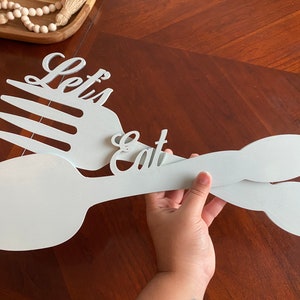 May include: White wooden cutout of a fork and spoon with the words "Let's Eat" on them.