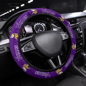 May include: Purple steering wheel cover with the word "Vikings" in white script and a Viking logo. The cover is on a black steering wheel inside a car.