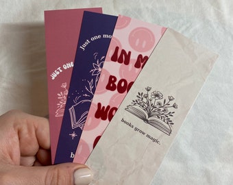 May include: Four rectangular bookmarks in a hand, featuring various designs and text. The bookmarks are in shades of pink, purple, and cream. Text includes "JUST ONE", "Just one more", "IN MY BOOK WORLD", and "books grow magic."