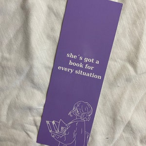 Könnte beinhalten: Lilafarbener Lesezeichen mit einer weißen Umrandung einer Person, die ein Buch liest. Der Text auf dem Lesezeichen lautet "she's got a book for every situation."