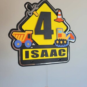 Puede incluir: Topper de pastel en forma de se&ntilde;al de construcci&oacute;n amarilla con un n&uacute;mero 4 negro, un cami&oacute;n volquete, una excavadora, una llave inglesa y un cono de tr&aacute;fico. El letrero tiene un borde negro con ribete plateado y el nombre "ISAAC" escrito en letras blancas.