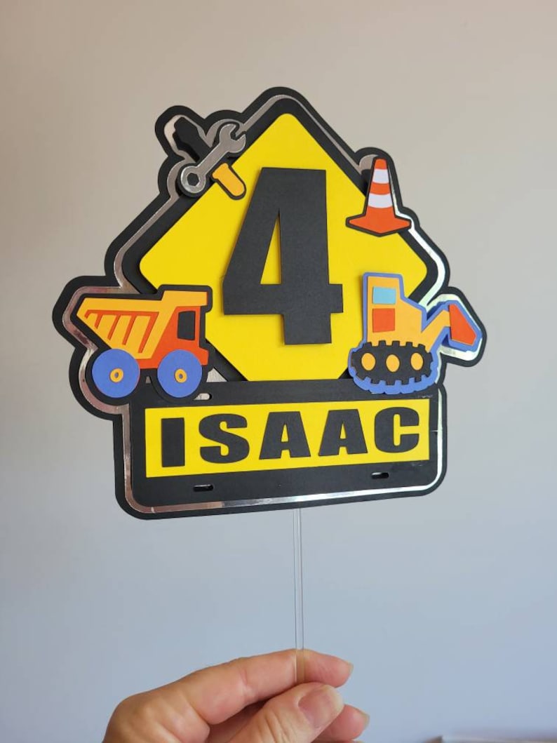 Puede incluir: Topper de pastel en forma de se&ntilde;al de construcci&oacute;n amarilla con un n&uacute;mero 4 negro, un cami&oacute;n volquete, una excavadora, una llave inglesa y un cono de tr&aacute;fico. El letrero tiene un borde negro con ribete plateado y el nombre "ISAAC" escrito en letras blancas.