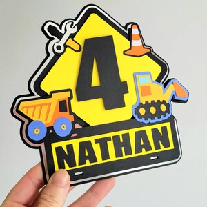 Puede incluir: Un cartel de construcci&oacute;n amarillo con un n&uacute;mero 4 negro, un cami&oacute;n volquete, una excavadora, una llave inglesa y un cono de tr&aacute;fico. El cartel tiene una placa negra con el nombre "NATHAN" en letras blancas.