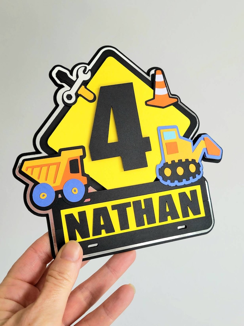 Puede incluir: Un cartel de construcci&oacute;n amarillo con un n&uacute;mero 4 negro, un cami&oacute;n volquete, una excavadora, una llave inglesa y un cono de tr&aacute;fico. El cartel tiene una placa negra con el nombre "NATHAN" en letras blancas.