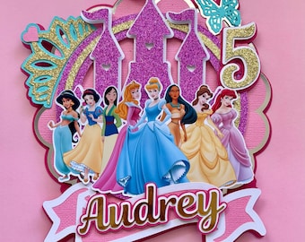 May include: A pink and gold cake topper with a castle, a butterfly, and the number 5. It features illustrations of princesses and the name "Audrey" in gold script. The topper is designed for a birthday celebration.