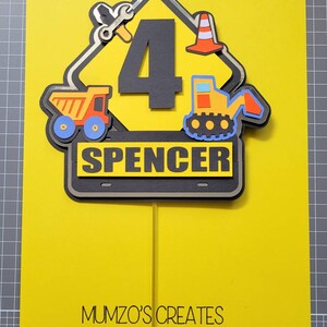 Puede incluir: Un topper de pastel de se&ntilde;al de construcci&oacute;n amarilla con un n&uacute;mero 4 negro, un cami&oacute;n volquete amarillo, una excavadora amarilla, una llave inglesa y un cono de tr&aacute;fico. El letrero tambi&eacute;n tiene el nombre "Spencer" escrito en &eacute;l. El topper de pastel est&aacute; sobre un fondo amarillo con el texto "MUMZO'S CREATES" en la parte inferior.