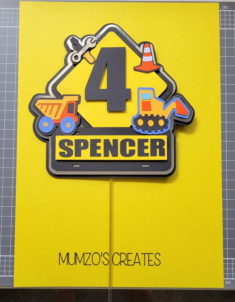 Puede incluir: Un topper de pastel de se&ntilde;al de construcci&oacute;n amarilla con un n&uacute;mero 4 negro, un cami&oacute;n volquete amarillo, una excavadora amarilla, una llave inglesa y un cono de tr&aacute;fico. El letrero tambi&eacute;n tiene el nombre "Spencer" escrito en &eacute;l. El topper de pastel est&aacute; sobre un fondo amarillo con el texto "MUMZO'S CREATES" en la parte inferior.