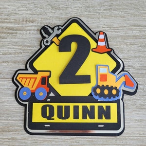 Puede incluir: Un cartel de construcci&oacute;n amarillo con un n&uacute;mero 2 negro, un cami&oacute;n volquete rojo y amarillo, una excavadora amarilla, una llave inglesa y un cono de tr&aacute;fico. El cartel tiene un borde negro y el nombre "QUINN" est&aacute; escrito en letras negras sobre una pancarta negra.