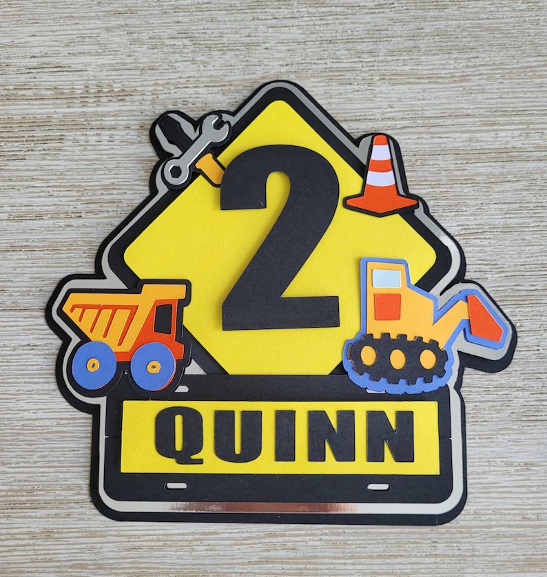Puede incluir: Un cartel de construcci&oacute;n amarillo con un n&uacute;mero 2 negro, un cami&oacute;n volquete rojo y amarillo, una excavadora amarilla, una llave inglesa y un cono de tr&aacute;fico. El cartel tiene un borde negro y el nombre "QUINN" est&aacute; escrito en letras negras sobre una pancarta negra.