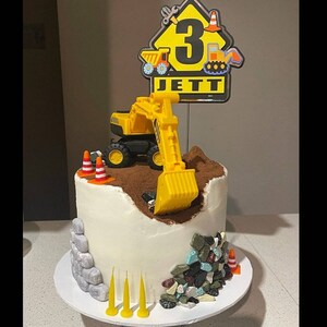 Puede incluir: Un pastel blanco decorado con un juguete de excavadora amarilla, tierra de chocolate y caramelos de roca de colores. El pastel est&aacute; cubierto con un letrero que dice "Jett" y "3" en texto negro.