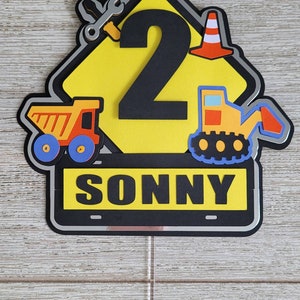 Puede incluir: Topper de pastel en forma de se&ntilde;al de construcci&oacute;n amarilla con un n&uacute;mero 2 negro, un cami&oacute;n de juguete, una excavadora de juguete, una llave inglesa y un cono de tr&aacute;fico. El letrero tambi&eacute;n tiene el nombre "SONNY" escrito en &eacute;l.