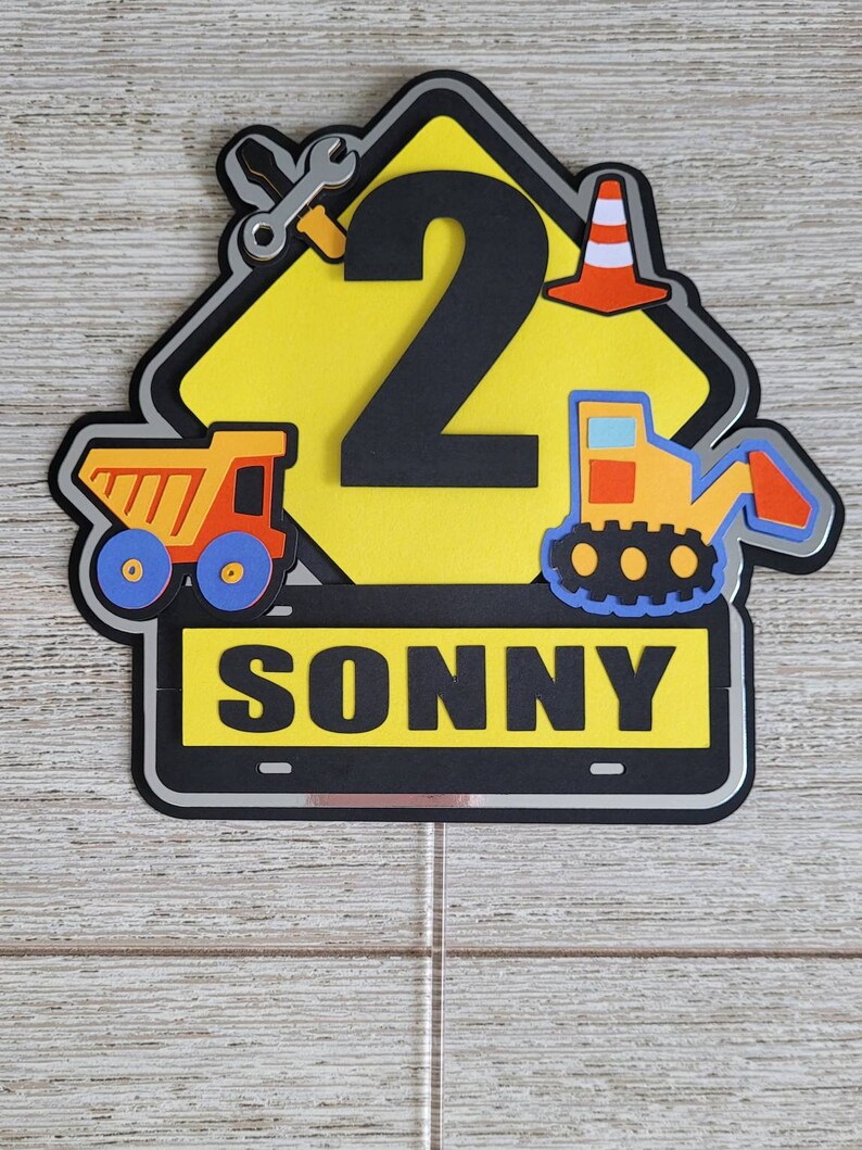 Puede incluir: Topper de pastel en forma de se&ntilde;al de construcci&oacute;n amarilla con un n&uacute;mero 2 negro, un cami&oacute;n de juguete, una excavadora de juguete, una llave inglesa y un cono de tr&aacute;fico. El letrero tambi&eacute;n tiene el nombre "SONNY" escrito en &eacute;l.
