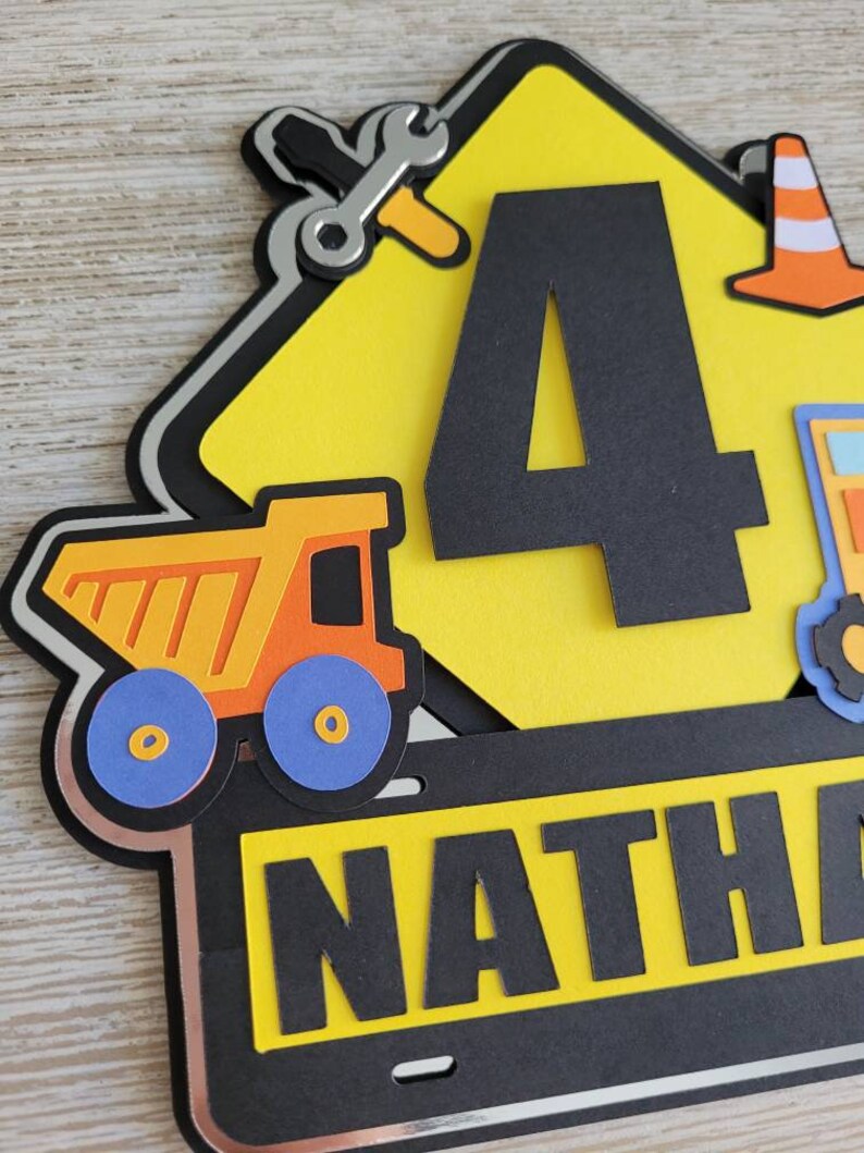 Puede incluir: Un cartel de construcci&oacute;n amarillo con un n&uacute;mero 4 negro, un cami&oacute;n volquete negro y amarillo, una llave inglesa y un destornillador negros, un cono de tr&aacute;fico negro y naranja y una placa de matr&iacute;cula negra con el nombre "NATHA" en letras negras.