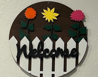 Letrero de bienvenida redondo de 30 cm, teñido en nogal, con valla blanca y detalles florales.