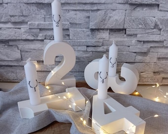 May include: White number-shaped candle holders with candles. The numbers 1, 2, 3, and 4 are displayed, each holding a white candle with a black antler design and a number. The candle holders are on a gray cloth with string lights.
