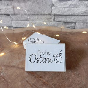 Könnte beinhalten: Zwei weiße rechteckige Schachteln mit den Worten "Frohe Ostern" in schwarzer Schrift und einem Hasengesicht. Die Schachteln befinden sich auf einer Holzoberfläche, mit Lichterketten im Hintergrund.