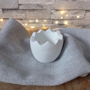 Könnte beinhalten: Ein weißer, eierschalenförmiger dekorativer Behälter mit gewelltem Rand. Der Behälter besteht aus einem glatten Material und liegt auf einem grauen Leinentuch. Der Hintergrund zeigt eine Backsteinmauer und Lichterketten.