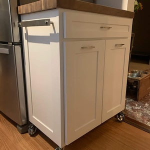 May include: White kitchen cabinet with a wood countertop and two doors. The cabinet has a drawer and is on wheels. The cabinet is in a kitchen with wood floors.