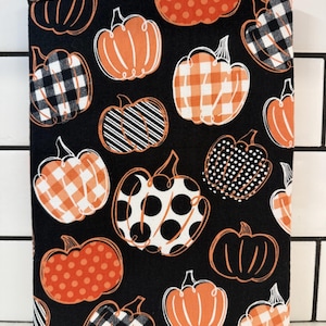 May include: A black fabric book sleeve featuring a repeating pattern of orange pumpkins in various designs, including polka dots, stripes, and plaid. The pumpkins are outlined in white, creating a festive fall theme.
