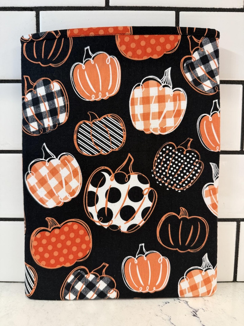 May include: A black fabric book sleeve featuring a repeating pattern of orange pumpkins in various designs, including polka dots, stripes, and plaid. The pumpkins are outlined in white, creating a festive fall theme.