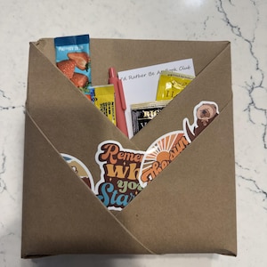 May include: A brown paper envelope containing a pink pen, a card with the text 'I'd Rather Be At Book Club', and several small packets. The front of the envelope is decorated with stickers, including one featuring an otter and the text 'the sun'.