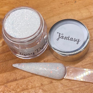 May include: A clear jar of white nail dip powder with iridescent glitter, next to a nail swatch and a lid labelled "Fantasy". The jar is open, revealing the powder's texture. The nail swatch displays the powder's application.