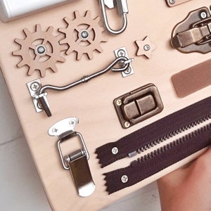 May include: A wooden board with various textures and activities for children. It features two gears, a star, a hook and eye latch, a latch, a zipper, and a spring-loaded clasp.