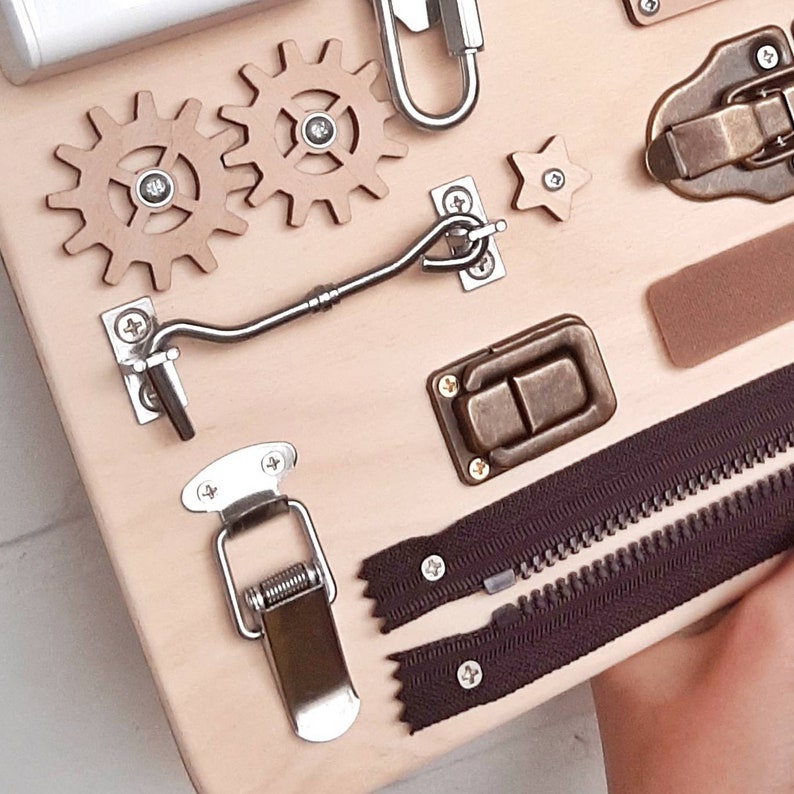 May include: A wooden board with various textures and activities for children. It features two gears, a star, a hook and eye latch, a latch, a zipper, and a spring-loaded clasp.