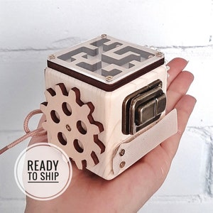 Può includere: Cubo di legno con un labirinto, un ingranaggio, un chiavistello e un cinturino in pelle. Il cubo è in legno chiaro con accenti marroni. Il cubo è tenuto in una mano.