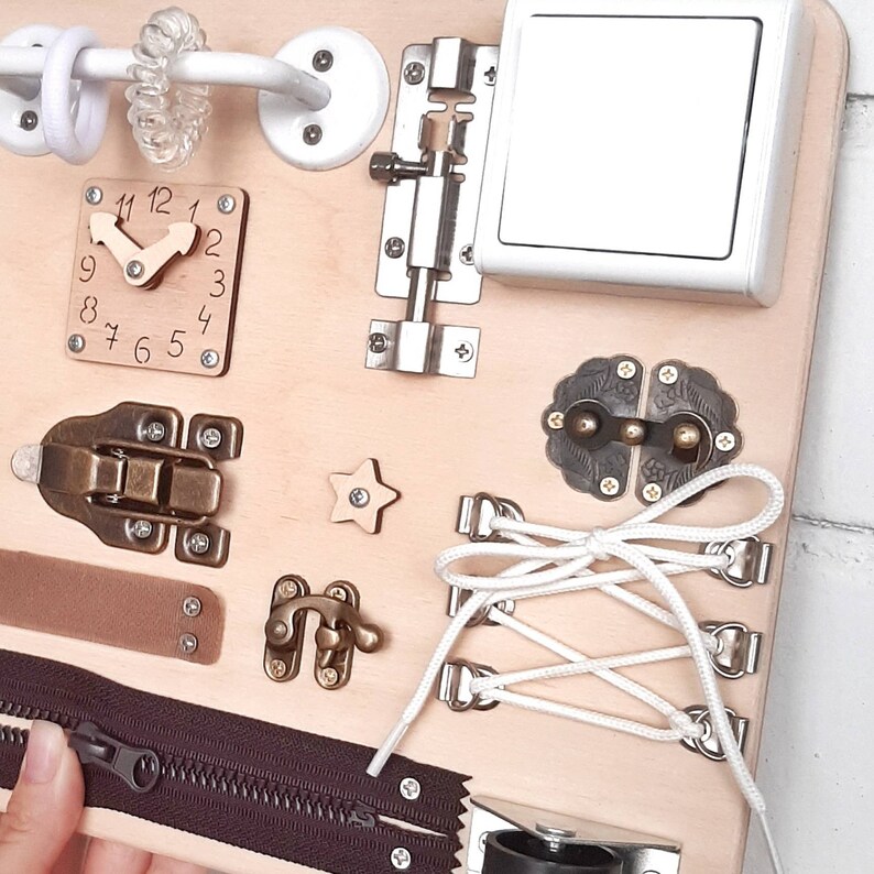May include: A wooden busy board with various activities for children, including a clock, a zipper, a latch, a star, a toggle, a buckle, a shoelace, and a light switch.