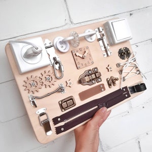 May include: A wooden activity board with various textures and activities for children. It features a zipper, a clock, a latch, a hook, a toggle switch, and gears. The board is light brown and has a natural wood finish.