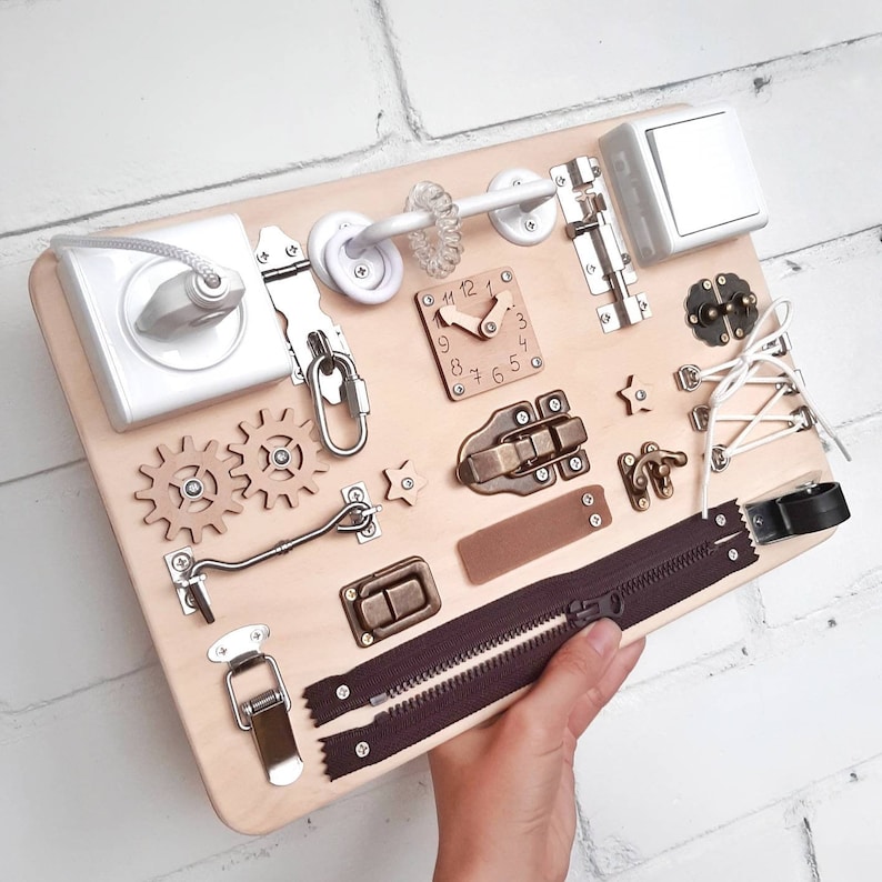 May include: A wooden activity board with various textures and activities for children. It features a zipper, a clock, a latch, a hook, a toggle switch, and gears. The board is light brown and has a natural wood finish.